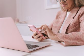 Mulher idosa usa um smartphone e um laptop sentada à mesa.
