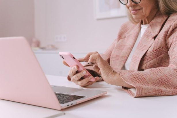 Mulher idosa usa um smartphone e um laptop sentada à mesa.